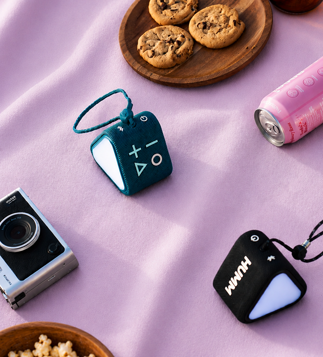 Two HUMM GLO Bluetooth speakers on a pink blanket with cookies and a camera in the background