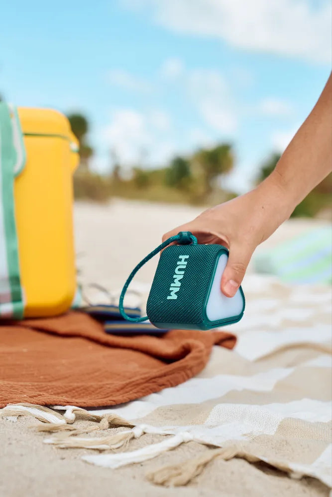 A hand picking up the waterproof HUMM GLO Bluetooth speaker from the picnic setting, including blankets and a yellow cooler. 