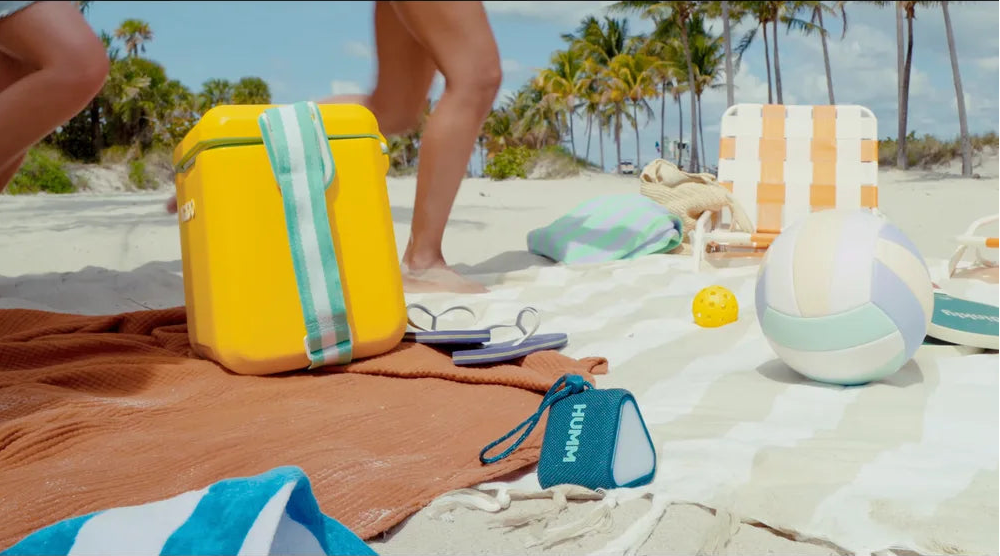 HUMM GLO Bluetooth speaker in a beach scene with a yellow bag, a blue pouch, and sunglasses on a towel with palm trees in the background.